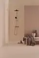 Duschsystem von Ideal Standard mit zwei Griffen in Chrom. Montiert an einer Wand, neben einer Ablage mit dekorativen Elementen und einem Handtuch. Minimalistisches Badezimmerdesign.